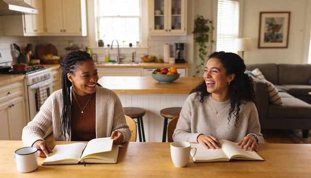 Morning journaling friends laughing at sunlit kitchen table with sweaters and notebooks
