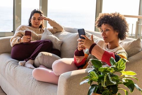 Multiracial women relaxing on sofa scrolling smartphones in bright cozy apartment living room