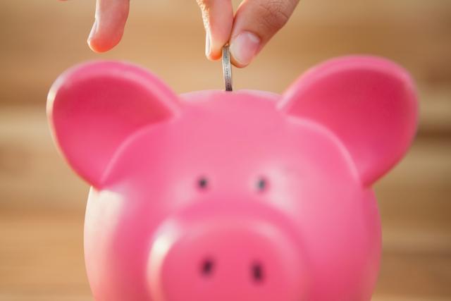 Close-up of a hand inserting a coin into a pink piggy bank. This image is ideal for illustrating concepts related to saving money, personal finance, budgeting, and financial planning. It can be used in articles, blogs, financial advice websites, and educational materials about money management and investment.