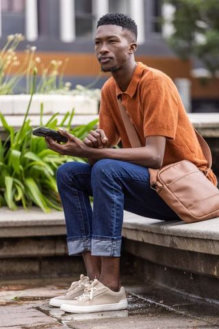 Young man sitting on urban bench holding smartphone wearing rust-orange polo and crossbody bag