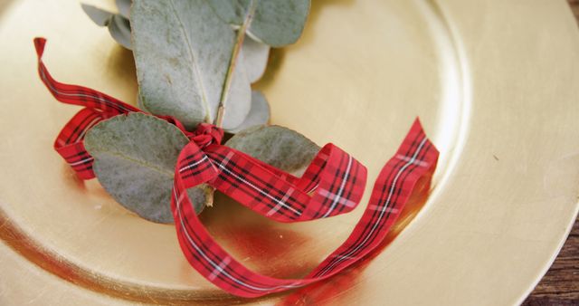 Eucalyptus leaves and a red plaid ribbon adorn a golden charger plate, creating a festive table setting. This arrangement suggests a holiday or special occasion celebration with an elegant touch.
