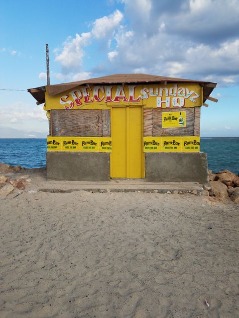 Small wooden bar hut with vibrant yellow door situated on a sandy beach overlooking the ocean. Ideal for use in travel brochures, vacation advertisements, tropical themed designs, or in articles about coastal destinations and beach bars.