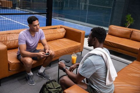 Male friends relaxing and talking in sports lounge by indoor court with phones and towel
