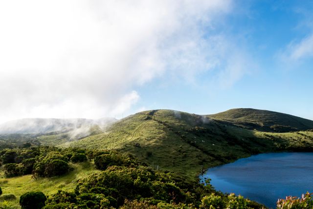 This image depicts lush green rolling hills partially covered in morning mist with a tranquil lake on the right side under a clear blue sky. It showcases the beauty and serenity of the natural landscape, making it ideal for use in travel brochures, nature documentaries, and inspirational posters promoting outdoor activities, relaxation, and the peacefulness of unspoiled environments.