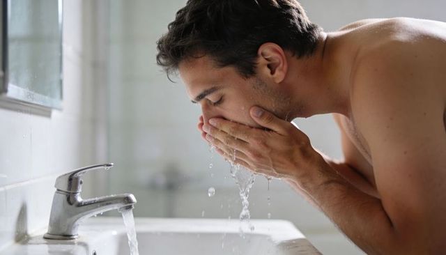 Shirtless man washing face at bathroom sink during morning grooming routine