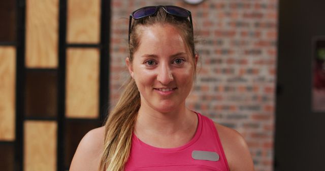 A woman with a light brown ponytail and wearing sunglasses on her head, smiling in a gym. She is in fitness attire, including a pink sleeveless shirt. The background features a brick wall and wooden panels. This can be used for fitness promotions, healthy lifestyle blogs, gym advertisements, and wellness content.