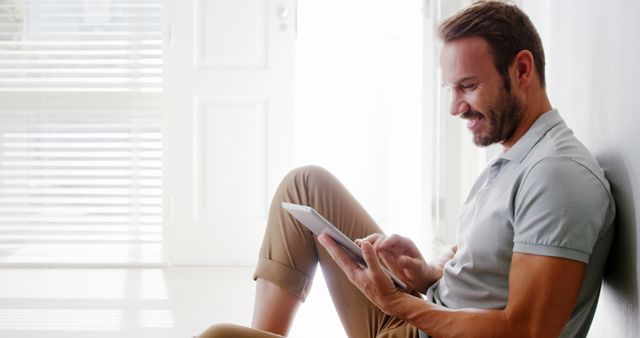 Young man sitting against wall using tablet and smiling. Casual attire, modern home setting with natural light. Perfect for themes promoting technology use, relaxed lifestyles, or work-from-home environments.