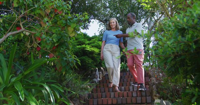 This stock photo shows a cheerful couple walking down steps surrounded by a lush garden filled with greenery. Perfect for articles or advertisements related to aging gracefully, spending time outdoors, senior relationships, or promoting a healthy and active lifestyle in older adults.