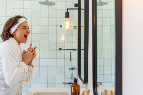 Laughing woman clasping hands at bathroom vanity wearing white robe and headband, morning routine