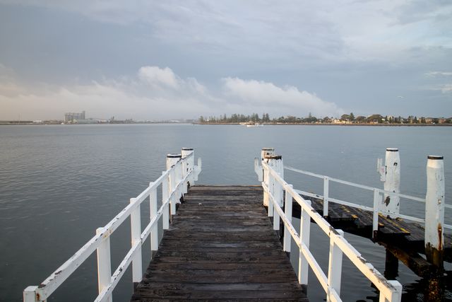 This image depicts an old wooden dock stretching over calm water with a cloudy sky in the background. The scene is tranquil and serene, making it perfect for illustrating concepts of peacefulness, relaxation, and nature. It can be used for travel and vacation promotions, website banners, or as a calming visual in various media.