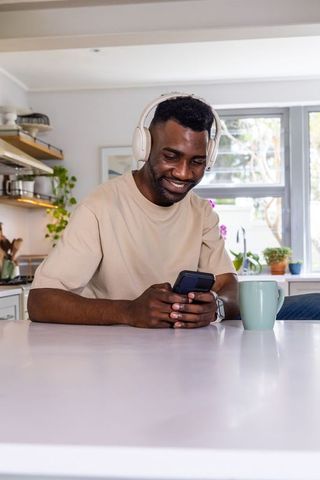 Smiling african man wearing wireless headphones using smartphone at modern kitchen island