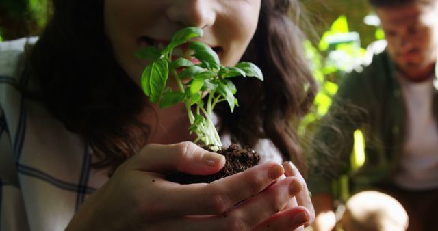 A young Caucasian girl gently smells a small plant she's holding, with copy space. Surrounding her, diverse individuals engage in gardening activities, emphasizing a connection with nature and environmental education.