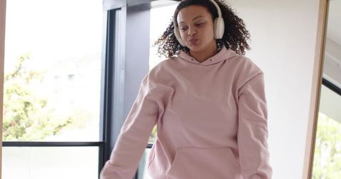 Curly-haired african american woman listening to music in pink hoodie by balcony window