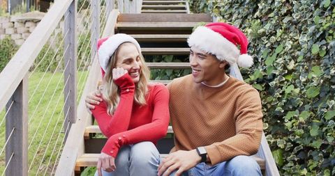 Couple wearing santa hats sitting on garden stairs sharing laughs and festive warmth