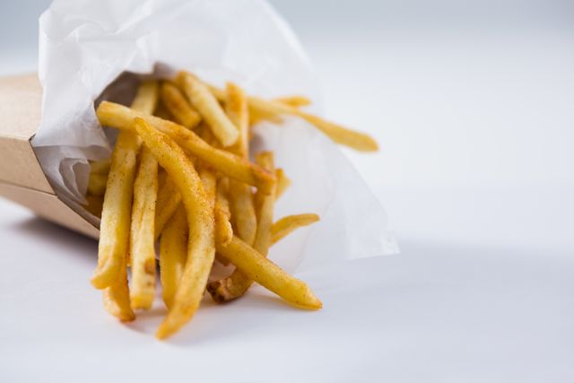 Golden, crispy french fries spilling out of a paper container against a clean white background. Perfect for illustrating fast food concepts, unhealthy eating habits, or takeout meal options. Ideal for use in advertisements, menus, or food blogs.