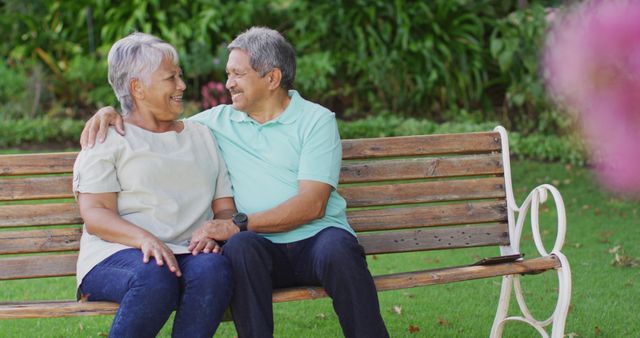 Senior couple smiling and bonding while sitting on a wooden bench in a lush garden. This image can be used in campaigns promoting senior health, retirement lifestyle, outdoor activities for elderly, and family values. Perfect for use in brochures, advertisements, and social media posts related to aging gracefully, senior community living, and family care.