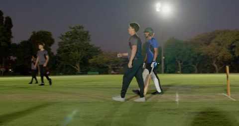 Evening cricket match under floodlights with bowler running and batsman questioning umpire