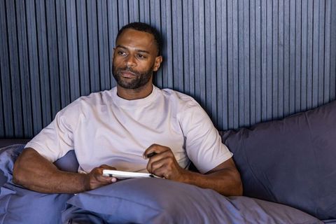Mature african american man relaxing in bed using tablet with stylus, contemplative modern bedroom