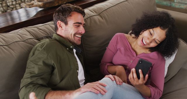 Biracial couple smiling while looking in smartphone together on the couch at vacation home. couple honeymoon and vacation concept