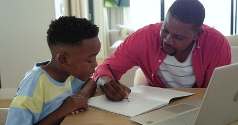 African american father tutoring son at home focusing on homework with laptop
