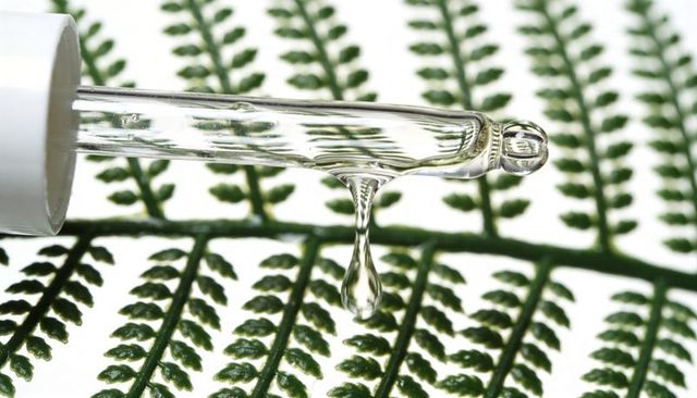 Glass dropper releasing clear serum over fern frond drip botanical skincare concept