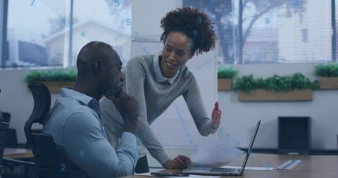 Woman guiding coworker through data report on laptop during office strategy meeting