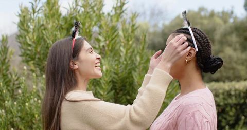 Smiling friends adjusting festive headbands in green garden wearing cozy sweaters
