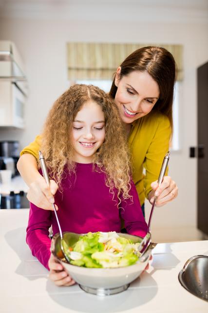 This image captures a tender moment between a mother and her daughter as they prepare a salad together in the kitchen. They are using fresh ingredients and sharing quality time, which emphasizes the themes of family bonding, healthy eating, and parenting. Ideal for use in articles or advertisements promoting family activities, healthy lifestyles, or cooking guides.