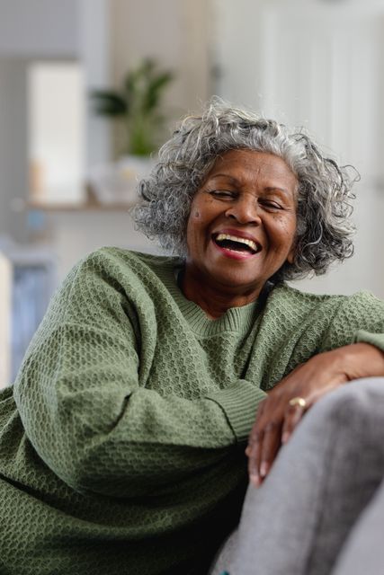 Vertical portrait of senior african american woman on sofa, laughing in living room, copy space. Retirement, relaxation, senior health, happiness, inclusivity and senior lifestyle concept.