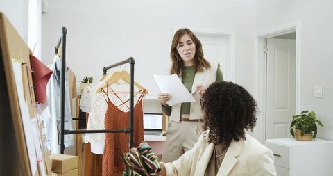 Diverse female designers discussing garment samples in bright studio with clothing rack