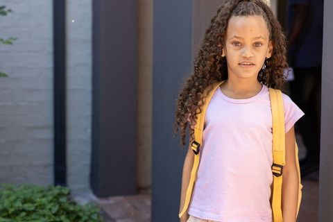 Curly-haired girl wearing yellow backpack standing at school entrance, back-to-school ready
