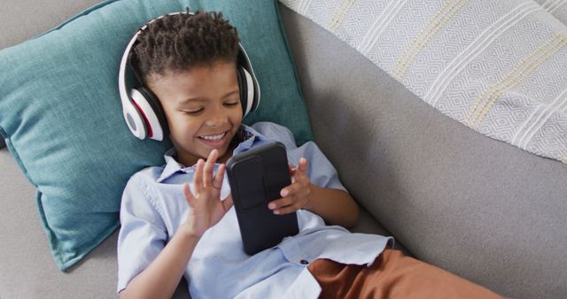 Boy with afro hair wearing headphones and using smartphone while lying on sofa, smiling in enjoyment. Suitable for articles on children's technology use, home entertainment, family living, or modern lifestyle. Could be used for promoting headphones, smartphones, or streaming services.