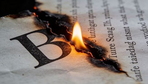 Burning newspaper closeup showing charred serif b, jagged tear and glowing embers