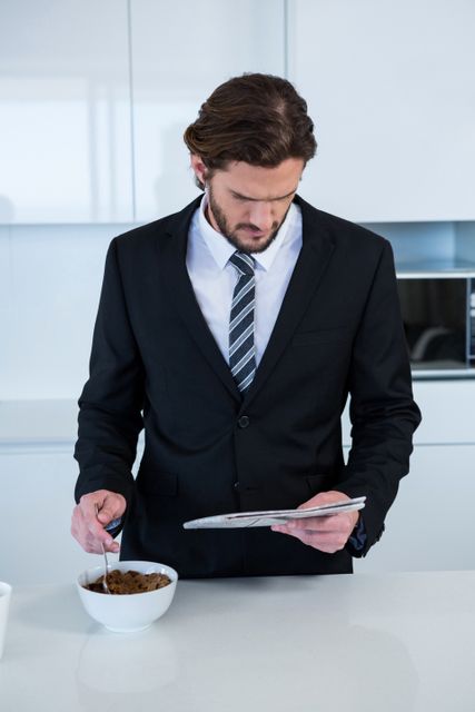 Businessman in a suit and tie reading a newspaper while having breakfast in a modern kitchen. Ideal for use in articles or advertisements about work-life balance, morning routines, professional lifestyles, or home office setups.