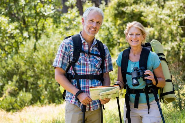 Senior couple enjoying a hike in a lush forest, holding a map and binoculars. Ideal for use in travel brochures, retirement lifestyle promotions, health and wellness campaigns, and outdoor adventure advertisements.
