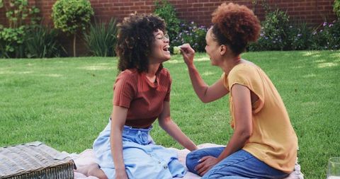 Two African American Women Sharing Grapes on Picnic Blanket in Sunny Backyard Garden