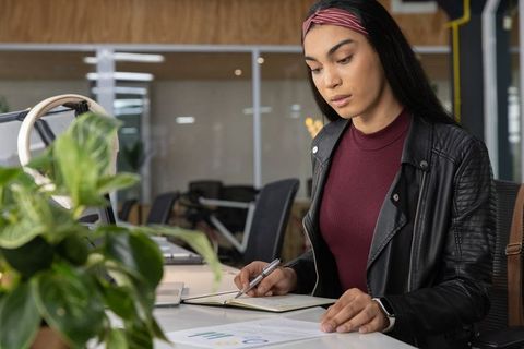 Young woman writing notes at open-plan office desk wearing leather jacket and smartwatch