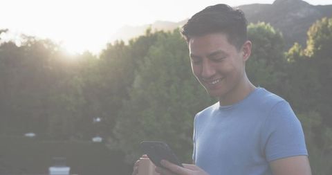 Smiling Asian man using smartphone on terrace with coffee cup at sunset, outdoor lifestyle