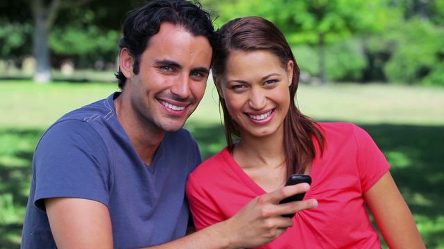 Couple enjoying time and sharing smiles while looking at mobile phone in tranquil park surroundings. Great for concepts related to technology, happiness, relationships, and outdoor activities. Ideal for websites, advertisements, or social media promoting lifestyle, mobile services, or tech engagement.