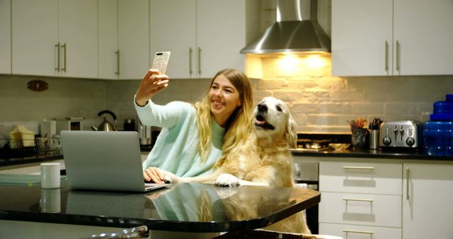 Young woman taking selfie with her dog in modern kitchen. They are sitting at a countertop with a laptop open, creating a mix of modern technology and warmth from the pet's companionship. Ideal for use in articles and content about digital lifestyles, home activities, pet ownership, and the joy of human-animal relationships.