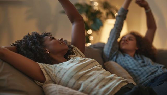 Two women relaxing and stretching on cozy living room sofa with warm string lights