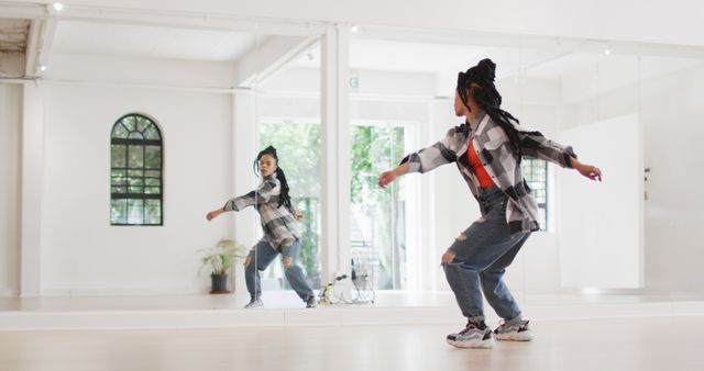 Young woman wearing a casual outfit with a flannel shirt and ripped jeans dancing hip hop in a bright studio. She is reflected in a large mirror, showing her dynamic movements. This image can be used to promote dance classes, fitness routines, or urban fashion.