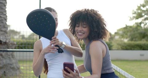 Smiling diverse women checking smartphone while holding paddle on outdoor paddle court