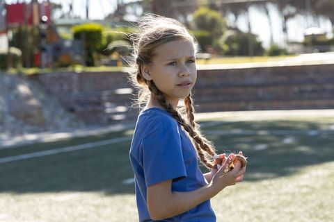 Backlit girl with braided pigtails holding dried grass on park turf at golden hour