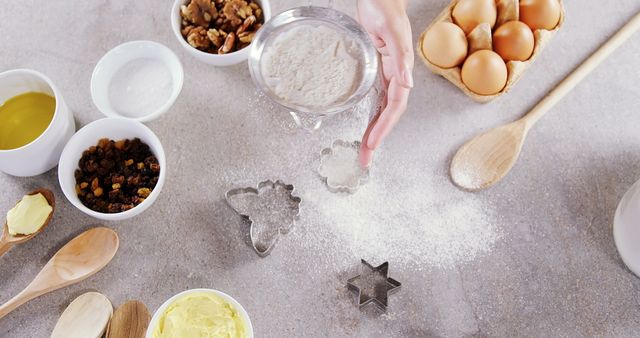 Person preparing dough with cookie cutters, surrounded by eggs, flour, butter, raisins, and walnuts. Use for cooking blogs, recipes, baking tutorials, and culinary websites.