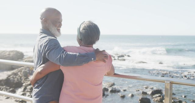 Senior couple smiling and holding each other on a beach promenade with the ocean in the background. Perfect for concepts related to love in later life, retirement, happiness, and enjoying the outdoors in older age.