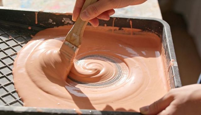 Holding wooden flat brush stirring peach paint spiral in black tray closeup