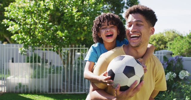 This image depicts a joyful father and son bonding while playing soccer in their backyard on a sunny day. The father holds a soccer ball and the son clings happily to his back, both smiling. The background shows lush greenery and a fence, indicative of a home garden or yard. Ideal for use in advertisements, blogs, or articles focusing on family, outdoor activities, and parent-child relationships.