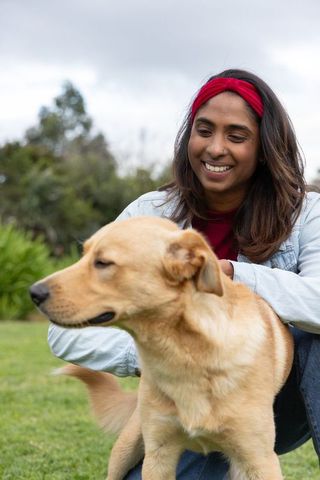 Indian woman smiling petting golden dog on park lawn wearing denim jacket and red headband