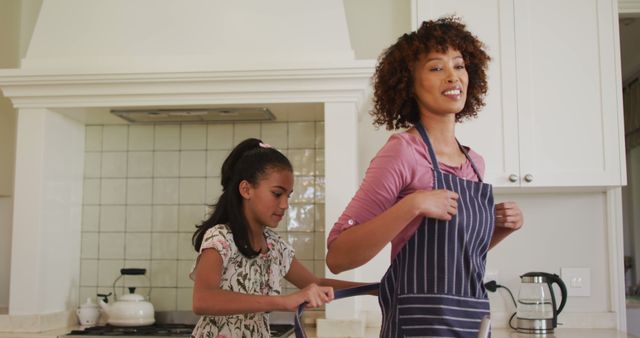 African american girl tying apron on her mother in the kitchen at home. family, love and togetherness concept